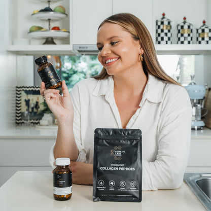 Smiling woman in a kitchen holding a bottle of Genetic Labs Australia supplement, with a package of Hydrolysed Collagen Peptides and another supplement bottle on the table in front of her.